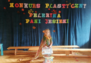 Dziewczynka w swojej pelerynce pozuje z nagrodami na tle napisu Konkurs Plastyczny Peleryna Pani Jesieni.