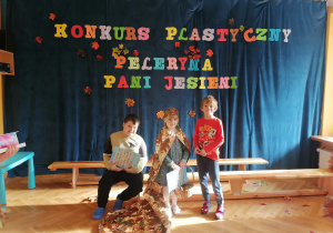 Dwóch chłopców i dziewczynka w swojej pelerynce pozują z nagrodami na tle napisu Konkurs Plastyczny Peleryna Pani Jesieni.