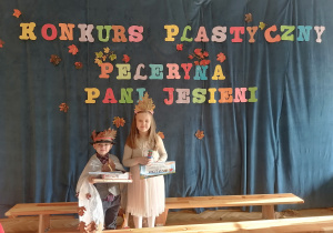 Chłopiec i dziewczynka w swoich pelerynkach pozują z nagrodami na tle napisu Konkurs Plastyczny Peleryna Pani Jesieni.