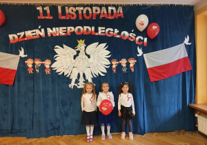 Trzy dziewczynki pozują na tle orła białego, polskich flag oraz napisu 11 listopada Dzień Niepodległości.