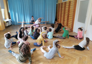 Dzieci siedzą na podłodze i wspólnie z trenerką zumby wykonują ćwiczenia rozciągające, tj. skłony w bok.