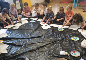 Dzieci siedzą na podłodze w kole i malują farbami plakatowymi krajobraz.
