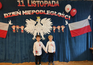 Dwóch chłopców pozuje na tle orła białego, polskich flag oraz napisu 11 listopada Dzień Niepodległości.