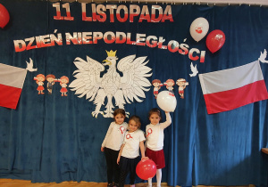 Trzy dziewczynki pozują na tle orła białego, polskich flag oraz napisu 11 listopada Dzień Niepodległości.