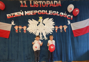Dwóch chłopców z kciukami uniesionymi do góry pozuje na tle orła białego, polskich flag oraz napisu 11 listopada Dzień Niepodległości.