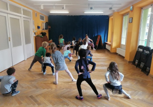 Dzieci stoją w rozsypance na sali i uczą się nowego układu tanecznego pokazywanego przez trenerkę zumby.