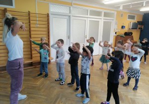 Dzieci stoją w rozsypance na sali i uczą się nowego układu tanecznego pokazywanego przez trenerkę zumby.