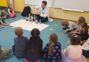 Dzieci siedzą w kole i obserwują doświadczenie chemiczne, które wykonuje Pan prowadzący na zajęciach Labolo.