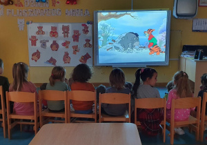 Dzieci siedzą na krzesełkach i oglądają bajkę " Kubuś Puchatek".
