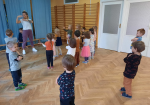 Dzieci stoją w rozsypce na sali i uczą się nowych ruchów do układu tanecznego.