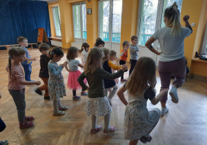 Dzieci stoją w rozsypance na sali, unoszą rękę i nogę do góry, powtarzając za trenerką ruchy.