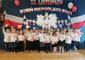 Dzieci stoją na tle orła białego, polskich flag oraz napisu 11 listopada Święto Niepodległości machając flagami wykonanymi własnoręcznie.