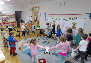 Dzieci tańczą w kole na dywanie ze swoimi pluszakami do piosenki o misiach.