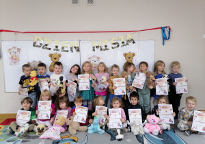 Dzieci ustawione w dwóch rzędach prezentują swoje pluszowe zabawki i dyplomy przyjaciela pluszowego misia.