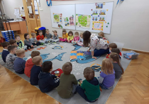 Dzieci siedzą w kole, a prowadzącą warsztaty rozkłada puzzle które będą podkładką do poruszania się dla robocika.