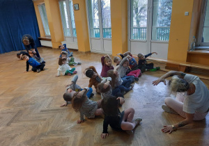 Dzieci siedzą na podłodze w siadzie skrzyżnym i wspólnie z trenerką wykonują skłony w bok. zumby wykonują