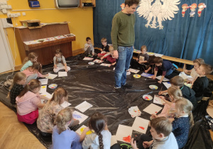 Dzieci siedzą w kole, przed nimi kartki i farby. Malują obraz inspirowany kierunkiem w malarstwie - impresjonizm.