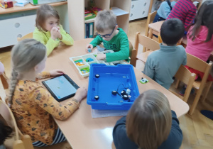 Dwie dziewczynki i dwóch chłopców siedzi przy stoliku i próbuje zbudować konstrukcje z klocków LEGO na zajęciach z robotyki z pomocą instrukcji wyświetlanej na tabelcie..