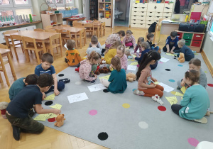 Dzieci siedzą na dywanie i podzielone na małe grupki układają postać misia z puzzli.