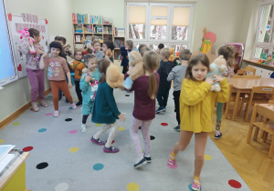 Dzieci tańczą do muzyki trzymając swoje misie na ramionach.