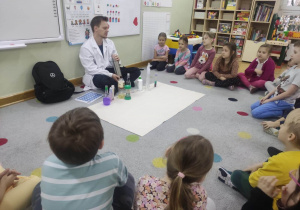 Dzieci siedzą w półkolu na dywanie i obserwują, jak prowadzący warsztaty wykonuje doświadczenie chemiczne.