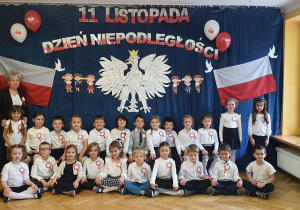 Dzieci wspólnie z Panią pozują na tle orła białego, polskich flag oraz napisu 11 listopada Dzień Niepodległości.