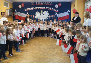 Dzieci stoją ustawione w półkolu, wspólnie z Paniami śpiewają Hymn Narodowy "Mazurka Dąbrowskiego" w rączkach trzymając biało - czerwone flagi Polski.