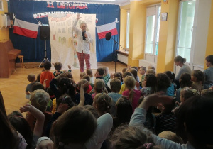Aktor występujący w przedstawieniu śpiewa piosenkę i wspólnie z dziećmi podnosi ręce wysoko do góry.
