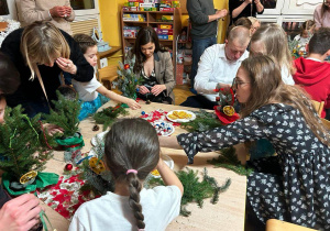 Warsztaty świąteczne. Dzieci siedzą przy stolikach i wspólnie z rodzicami wykonują stroiki świąteczne z gałązek choinki, bombek, wstążek, szyszek oraz suszonych pomarańczy z goździkiem podczas warsztatów świątecznych.
