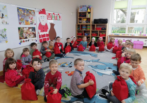 Dzieci siedzą w kole na dywanie prezentując swoje upominki od Świętego Mikołaja.
