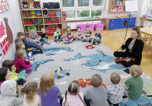 Dzieci siedzą na dywanie w kole i przyglądają się jak Pani logopeda prezentuje im smyczek od skrzypiec.