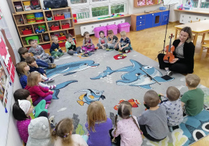 Dzieci siedzą na dywanie w kole i przyglądają się jak Pani logopeda prezentuje im skrzypce.