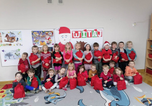 Dzieci ustawione w dwóch rzędach siedzą i stoją przy dekoracji i prezentują swoje upominki od Świętego Mikołaja.
