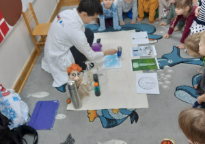 Dzieci siedzą w półkolu i obserwują, jak prowadzący wykonuje eksperyment polegający na zmianie koloru chmurki z obrazka.