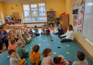 Akcja "Czytamy, głowy otwieramy". Dzieci siedzą w półkolu, a mama jednego z dzieci czyta im opowiadanie, a chłopiec pokazuje reszcie obrazki.