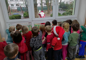 Dzieci stoją przy oknie i machają Świętemu Mikołajowi, który odwiedził przedszkole.