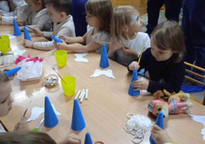 Dzieci siedzą przy stolikach i z pomocą rodziców wykonują własnego aniołka ze styropianowego stożka, pomalowanego na niebiesko, piórek, cekinów i innych materiałów.