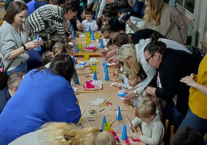 Dzieci siedzą przy stolikach i z pomocą rodziców wykonują własnego aniołka ze styropianowego stożka, pomalowanego na niebiesko, piórek, cekinów i innych materiałów.