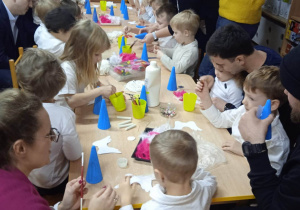 Dzieci siedzą przy stolikach i z pomocą rodziców wykonują własnego aniołka ze styropianowego stożka, pomalowanego na niebiesko, piórek, cekinów i innych materiałów.
