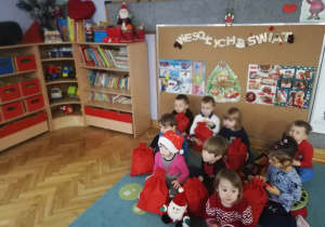 Dzieci siedzą na dywanie i prezentują upominki od Świętego Mikołaja.