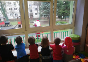 Dzieci stoją przy oknie i machają Świętemu Mikołajowi, który odwiedził przedszkole.