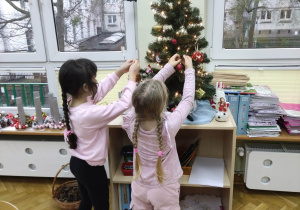 Dwie dziewczynki dekorują choinkę zawieszając bombki.