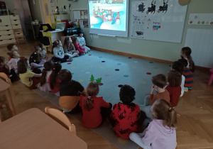 Dzieci siedzą w półkole na dywanie oglądając zimową bajkę na ekranie.