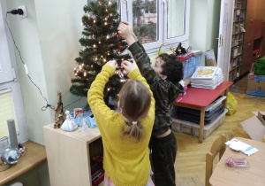 Chłopiec i dziewczynka dekorują choinkę zawieszając bombki.