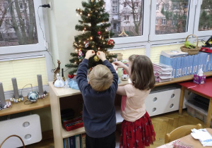 Chłopiec i dziewczynka dekorują choinkę zawieszając bombki.
