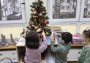 Dwie dziewczynki i chłopiec dekorują choinkę zawieszając bombki.