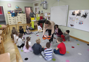 Dzieci siedzą na dywanie w kole. Nauczyciel logopeda trzyma w ręce skrzypce prezentując dzieciom instrument.
