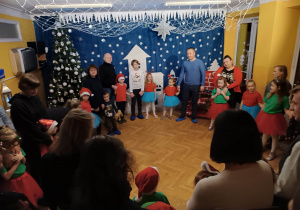 Dzieci stoją ustawione w kole i wspólnie z rodzicami śpiewają kolędy podczas występu świątecznego.
