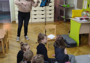 Nauczyciel trzyma w dłoniach skrzypce, dzieci siedzą na dywanie słuchając z zainteresowaniem kolęd.