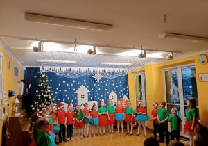 Dzieci stoją ustawione w półkolu i śpiewają piosenki podczas występu świątecznego.